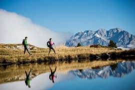 Zwei Nordic Walking Wandernde an einem Seeufer, dahinter eine Wolkenbank und schroffe Berge
