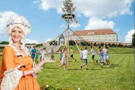 Im Vordergrund eine Dame in barockem Outfit die auf Jugendliche deutet, die um einen Maibaum tanzen, im Hintergrund Schloss Hof