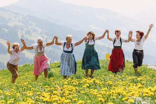 Sechs Damen in bunten Dirndls in einer L�wenzahnwiese, im Hintergrund die Alpen