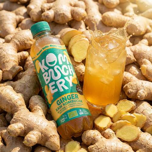 Eine Flasche Kombuchery Ginger neben einem engeschenkten Glas, im Hintergrund viele Ingwerknollen
